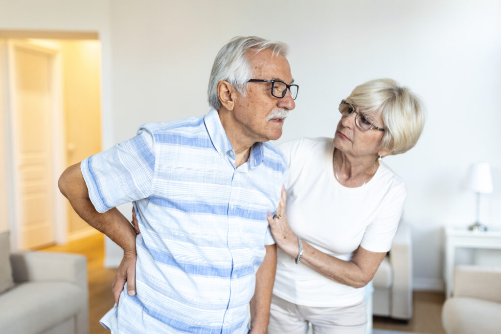 Elderly man experiencing lower back pain while being supported by his spouse at home