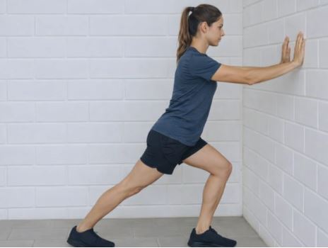 Woman performing a wall calf stretch with one leg stepped back, keeping the heel on the floor to stretch the calf and plantar fascia.